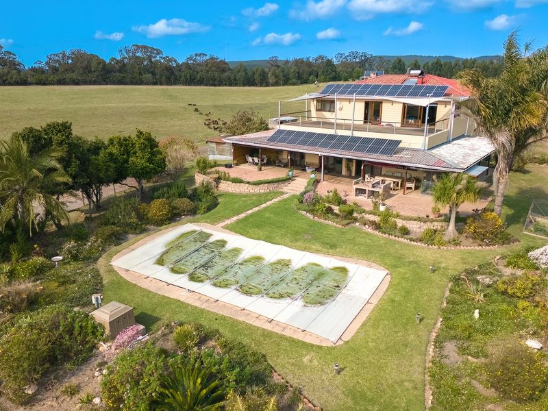 Aerial view of a farm property featuring a multi-level residence with solar panels, a swimming pool with cover, and expansive surrounding pasture land.