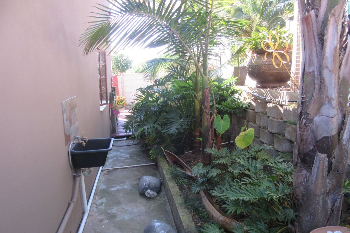 A narrow rear exterior side passage featuring a utility sink, concrete pathway, and a tiered retaining wall with established garden landscaping.