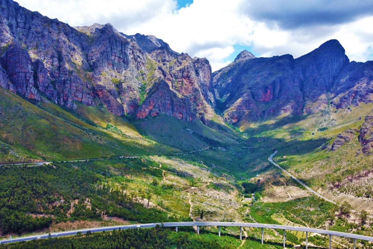 A scenic aerial view of a dramatic mountain range and valley landscape featuring a prominent highway bridge.