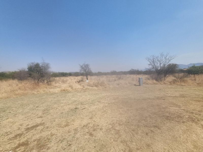 A wide-angle view of a vacant residential plot featuring dry grass, scattered indigenous trees, and a clear blue sky.