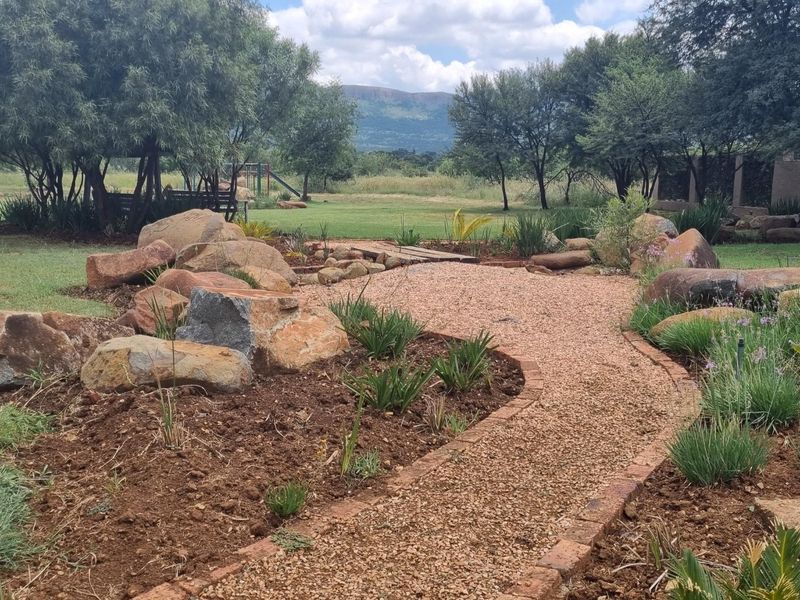 A landscaped garden area featuring a gravel pathway, decorative rockery, and mature trees with scenic mountain views in the background.