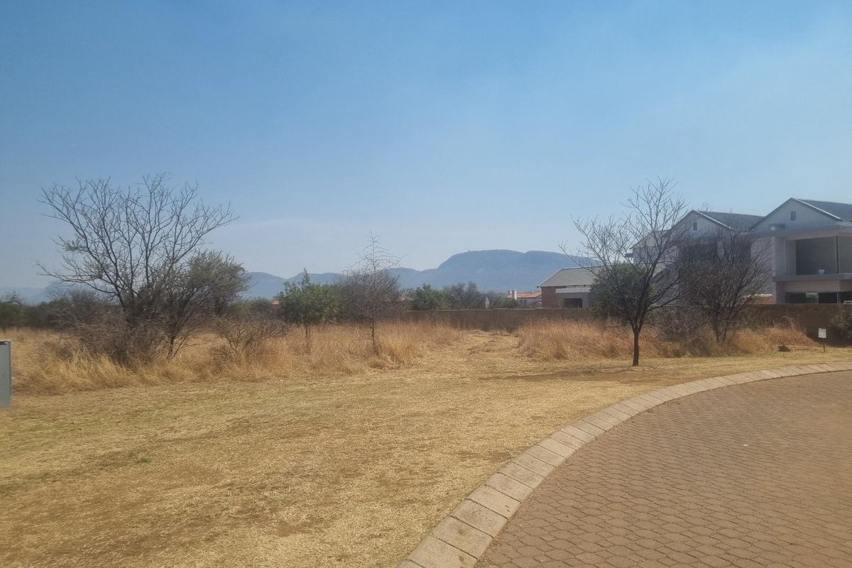 A vacant residential plot featuring dry grass, scattered trees, and mountain views, situated adjacent to a paved road in a residential estate.