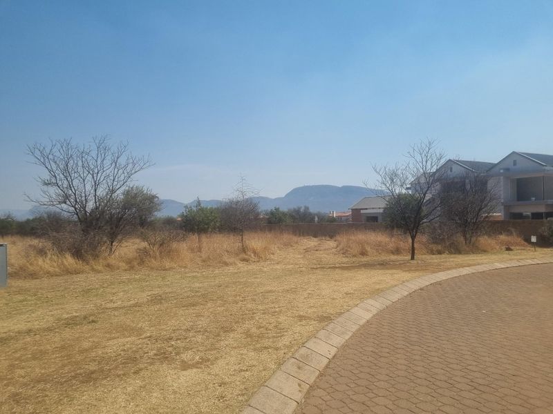 A vacant residential plot featuring dry grass, scattered trees, and mountain views, situated adjacent to a paved road in a residential estate.