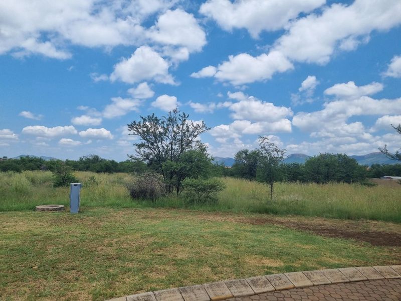 A view of a vacant residential plot featuring natural vegetation, a small utility pillar, and distant mountain views.
