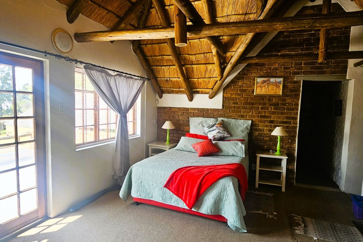 A rustic bedroom featuring exposed timber rafters, a traditional thatched roof, and an exposed brick feature wall with access to a private balcony.