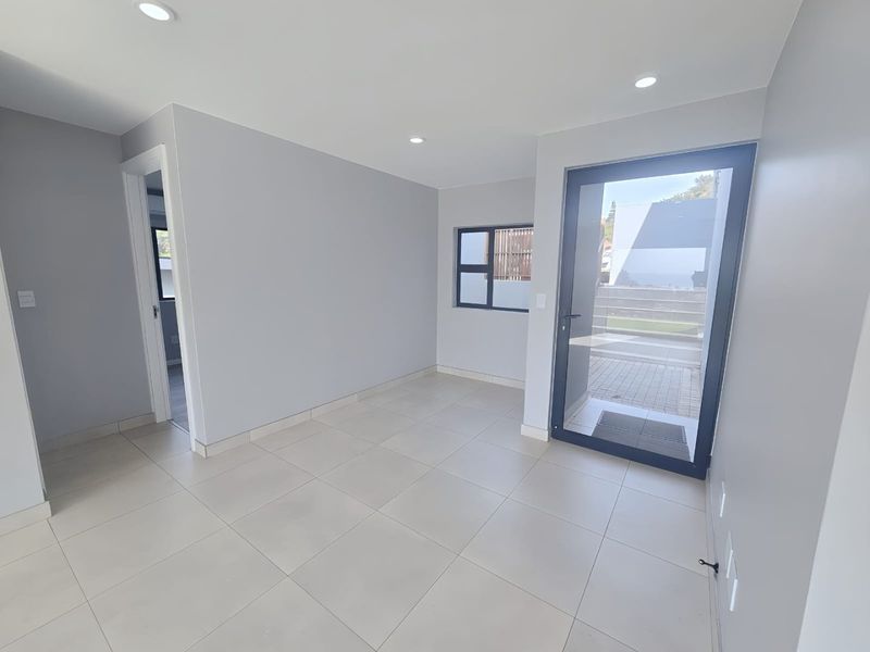 A bright, modern, and empty living area featuring neutral tiled flooring, recessed downlights, and direct access to an outdoor patio through a glass door.