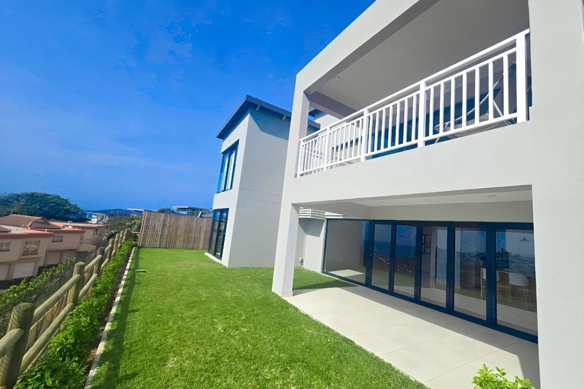 Rear view of a modern townhouse featuring a covered patio, upper balcony with white railings, large glass sliding doors, and a well-maintained lawn.