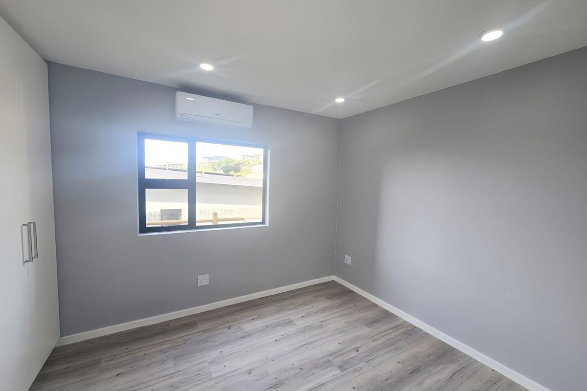 A clean, modern bedroom featuring neutral grey walls, durable laminate flooring, built-in wardrobe storage, and a wall-mounted air conditioning unit.