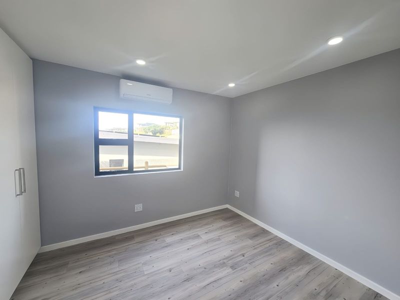 A clean, modern bedroom featuring neutral grey walls, durable laminate flooring, built-in wardrobe storage, and a wall-mounted air conditioning unit.