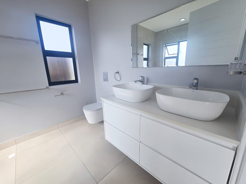 Modern bathroom featuring a sleek double vanity with vessel sinks, wall-mounted toilet, large mirror, and neutral floor tiling.