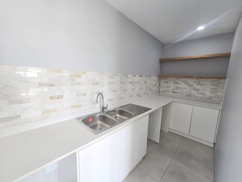 A clean, modern scullery featuring stone countertops, a double bowl stainless steel sink, tiled backsplash, and ample built-in cabinetry with floating timber shelving.