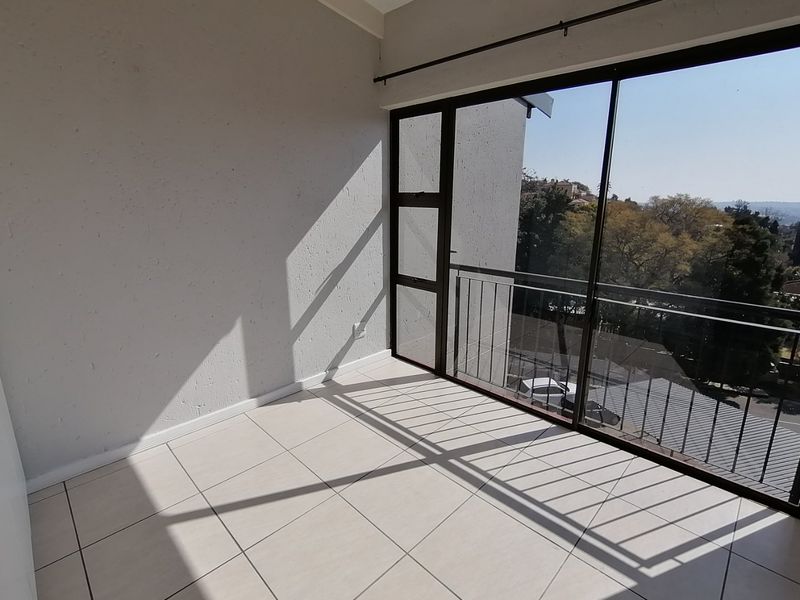 A bright, empty balcony featuring tiled flooring, a metal balustrade, and large sliding glass doors with views of the surrounding area.