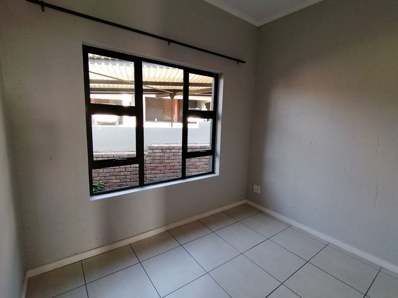 An empty bedroom featuring neutral tiled flooring, grey painted walls, and a black aluminium window frame overlooking the exterior area.