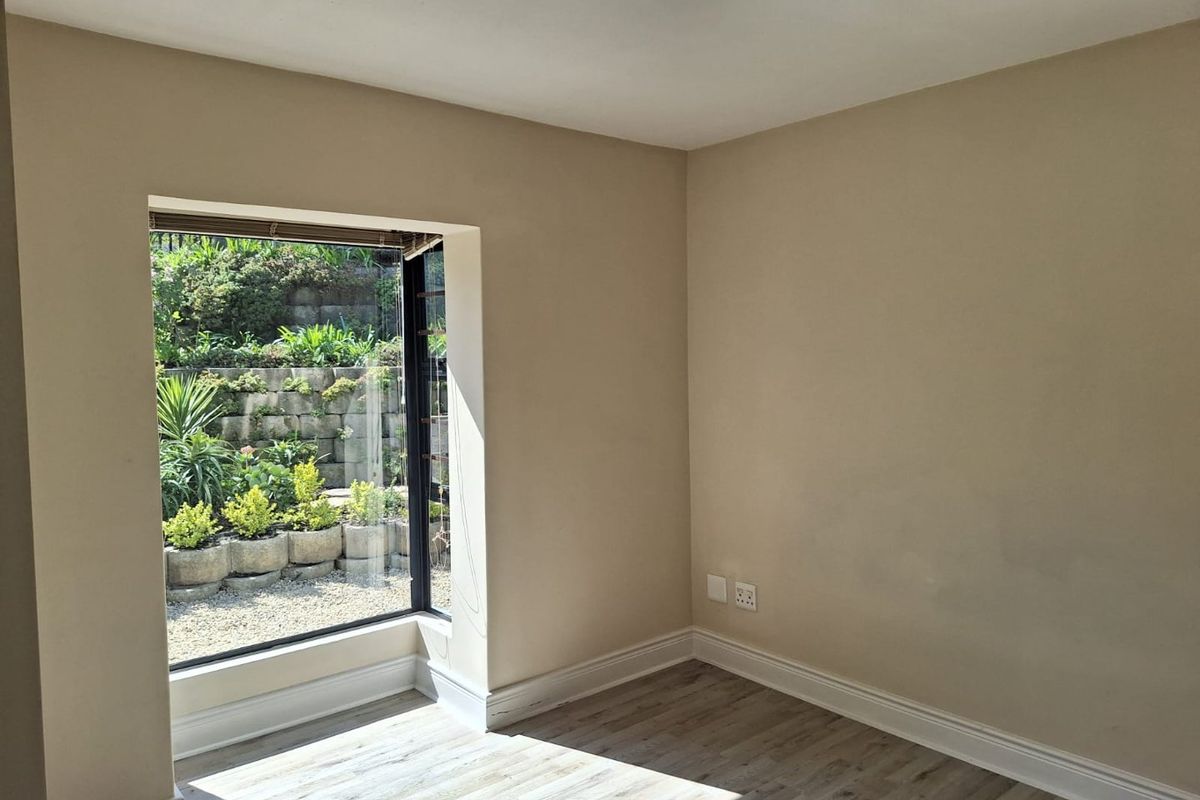 A bright, empty bedroom featuring modern laminate flooring, neutral walls, and a large window overlooking a landscaped garden.