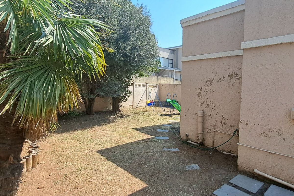 Rear garden area featuring a lawn, mature trees, boundary wall with electric fencing, and exterior wall requiring maintenance.