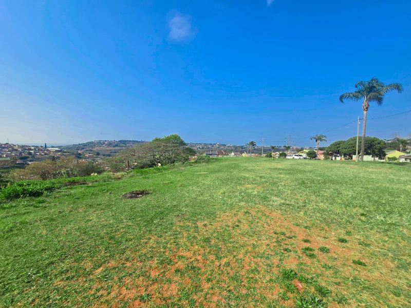 A wide-angle view of a vacant residential plot featuring open grassy terrain with distant neighborhood views and a clear blue sky.