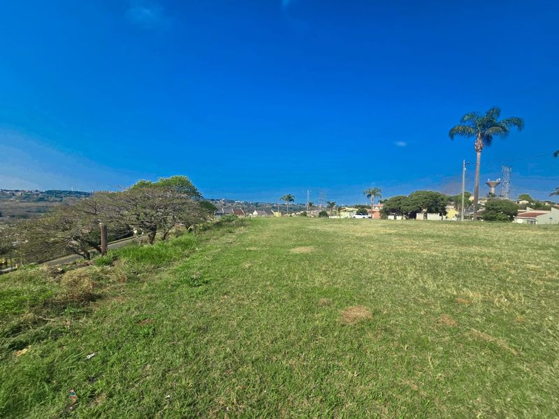 A spacious, open residential plot featuring established grass cover and mature trees, offering a clear view of the surrounding neighborhood.