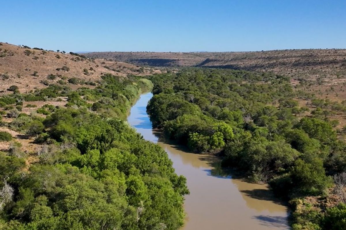 An aerial view of a natural river winding through the farm landscape, surrounded by lush green trees and arid hills.