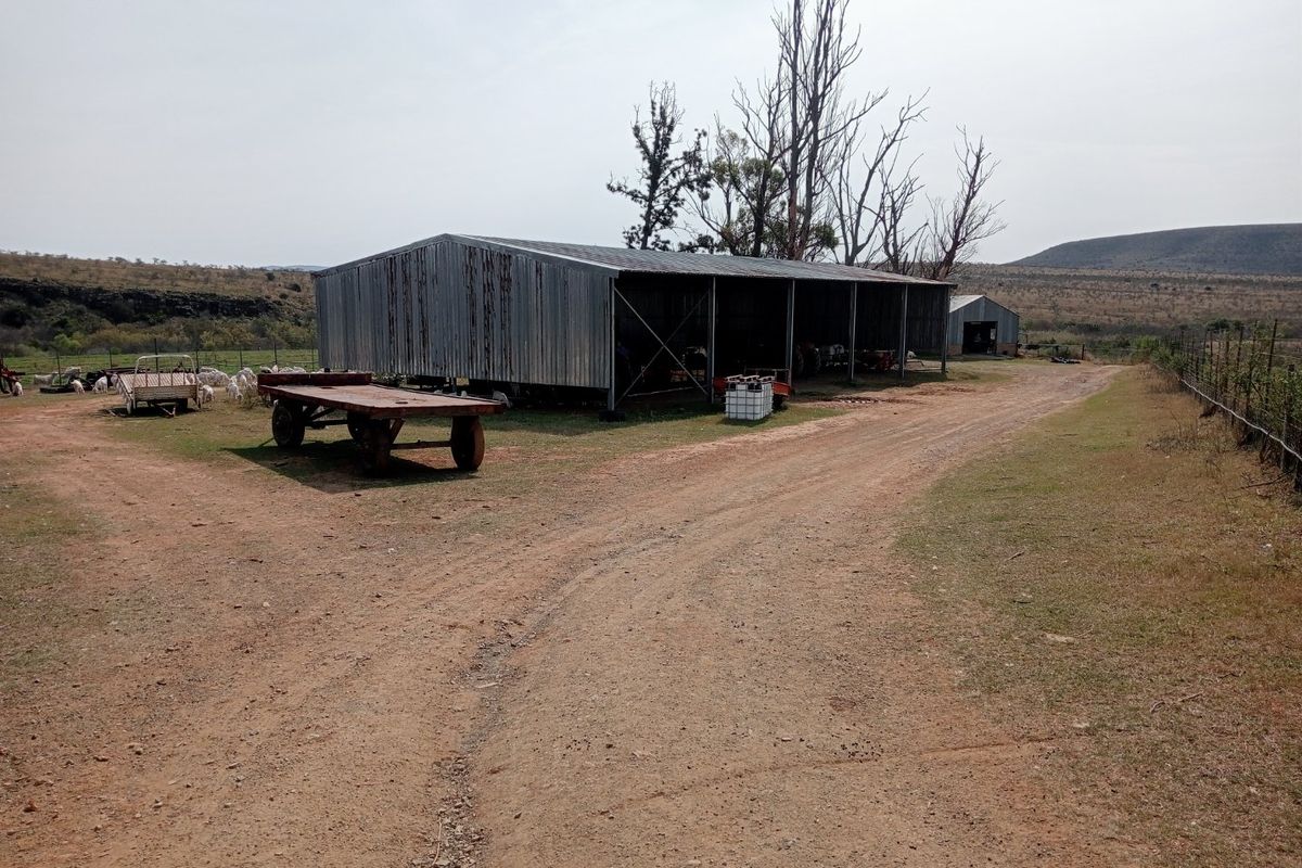 A functional farm yard featuring a large corrugated metal shed, dirt access paths, and surrounding agricultural fencing in a rural landscape setting.