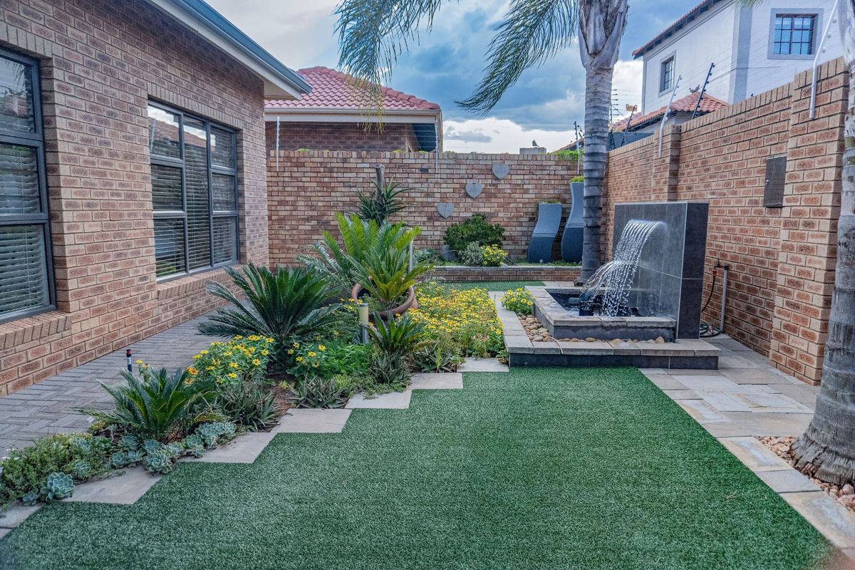 A well-maintained rear garden featuring a paved walkway, artificial lawn, and a modern stone water feature against a brick boundary wall.