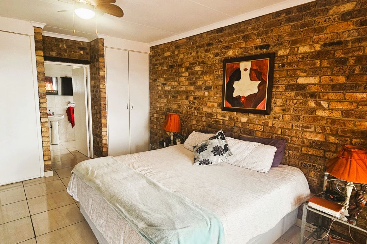 Spacious bedroom featuring an exposed brick accent wall, built-in storage cupboards, and tiled flooring, with an adjoining en-suite bathroom.