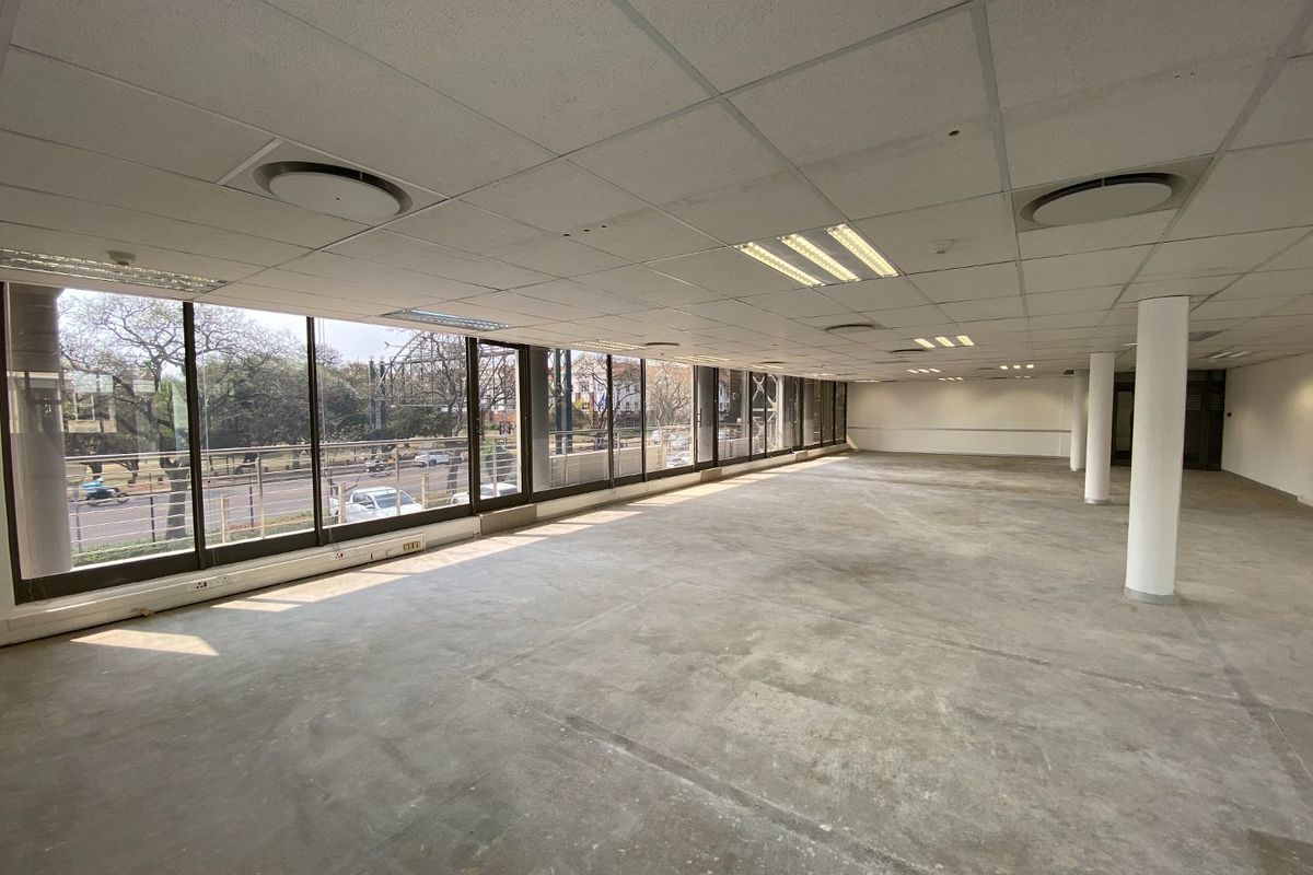 A spacious, open-plan commercial office area featuring large floor-to-ceiling windows, suspended ceilings with recessed lighting, and structural columns.