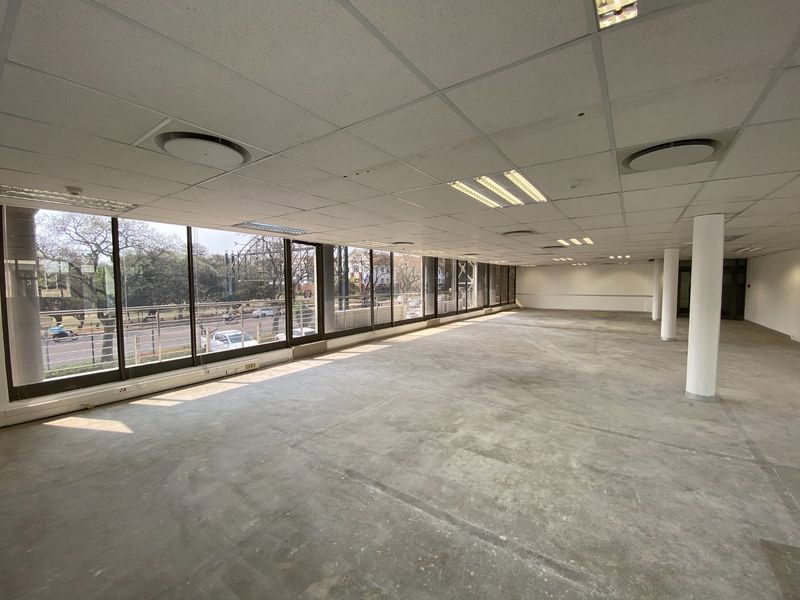A spacious, open-plan commercial office area featuring large floor-to-ceiling windows, suspended ceilings with recessed lighting, and structural columns.