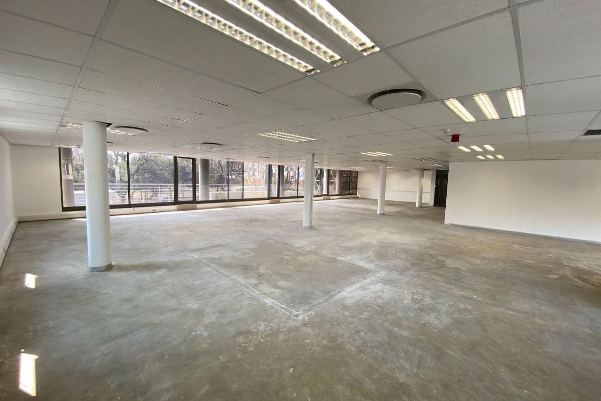 A large, open-plan commercial office space featuring structural columns, suspended ceilings with recessed lighting, and expansive windows providing natural light.