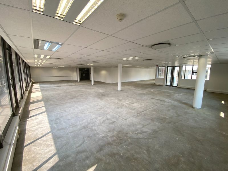 A large, open-plan commercial office space featuring a suspended ceiling, recessed lighting, structural columns, and expansive windows, currently awaiting floor finishing.