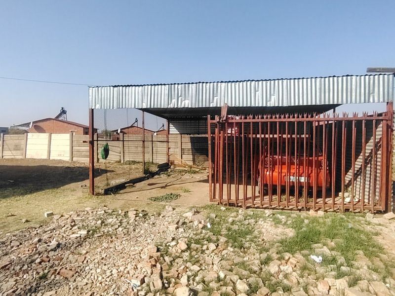 An outdoor carport structure featuring a corrugated metal roof and steel security gate, situated on an unpaved lot with perimeter boundary walls.