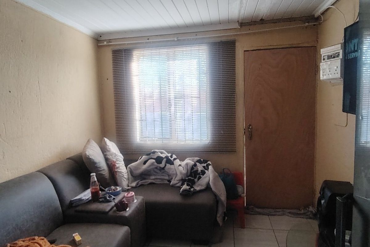 A compact living room featuring tiled flooring, a timber ceiling, and a window with venetian blinds, requiring cosmetic updates to improve its overall condition.