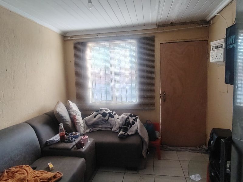 A compact living room featuring tiled flooring, a timber ceiling, and a window with venetian blinds, requiring cosmetic updates to improve its overall condition.