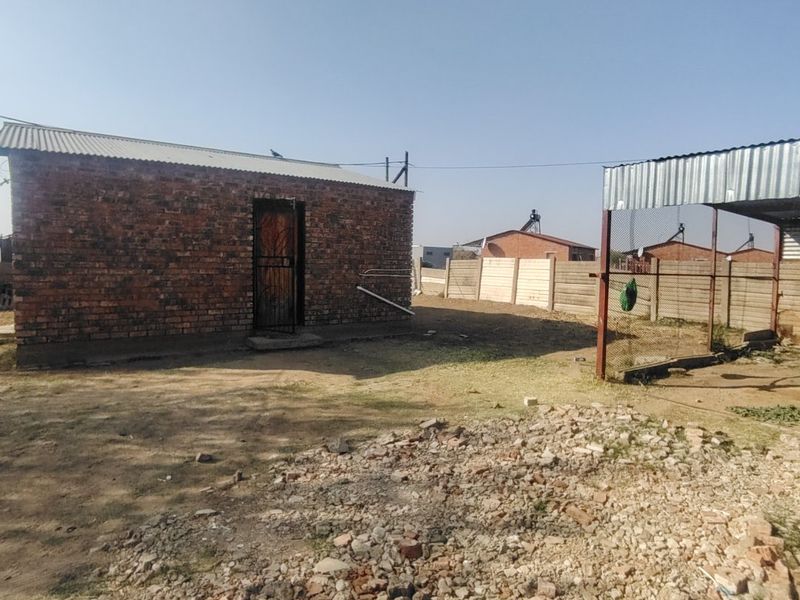 Rear exterior view of a face brick structure with a corrugated metal roof, featuring a fenced area and unpaved yard with loose rubble.