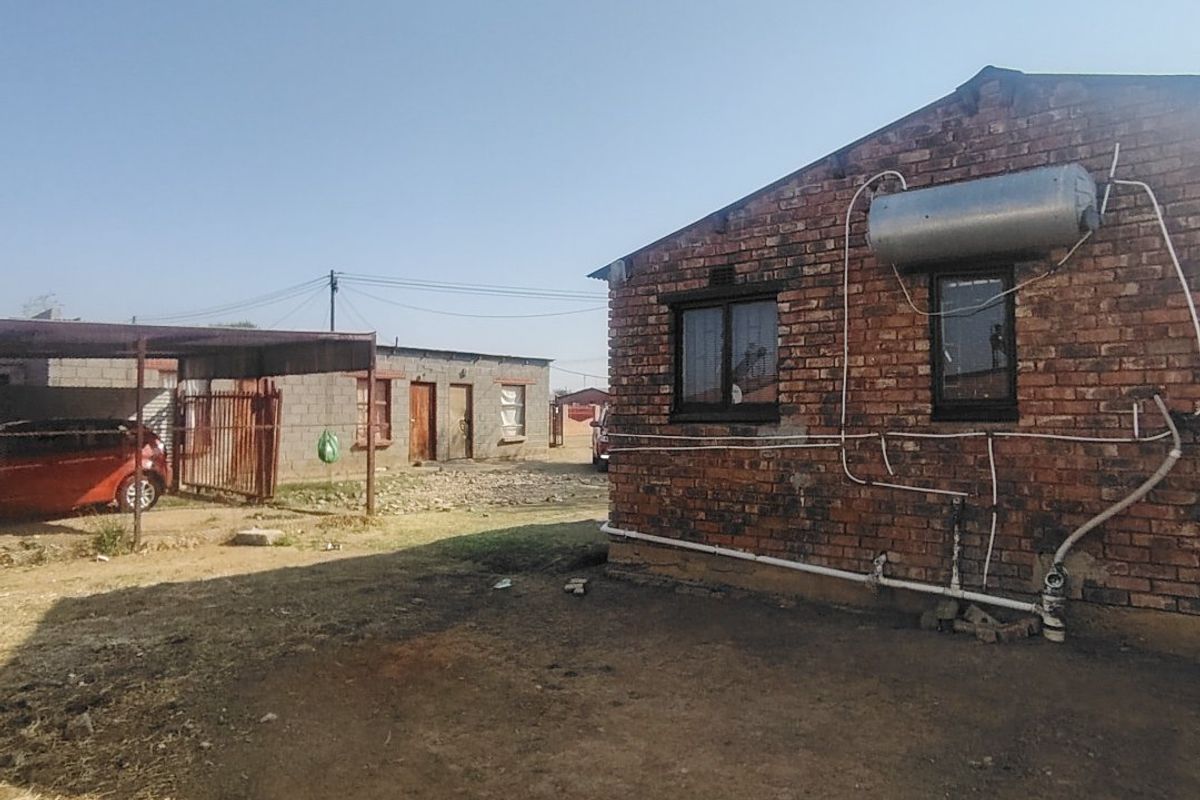 Rear view of a brick house featuring an external solar water heater, exposed plumbing, and an adjacent metal carport structure.