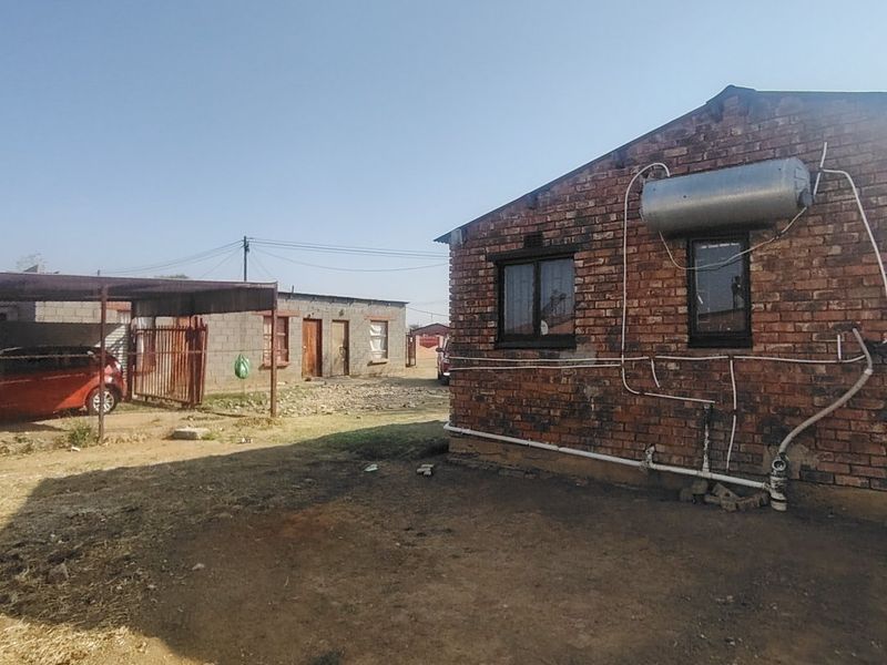 Rear view of a brick house featuring an external solar water heater, exposed plumbing, and an adjacent metal carport structure.