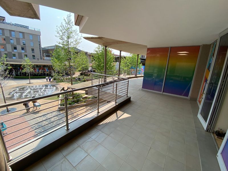 A spacious covered balcony featuring tiled flooring and a stainless steel balustrade, overlooking a commercial courtyard with a water feature.