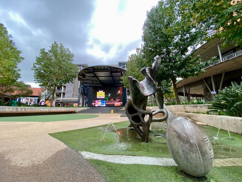 A well-maintained commercial outdoor plaza featuring a central sculpture, water feature, landscaped garden areas, and a stage with a large digital display.
