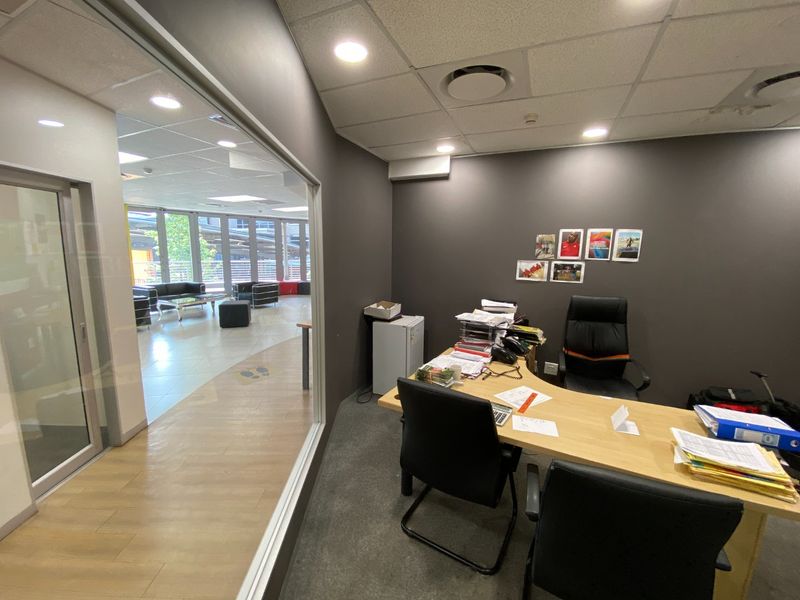 A professional office space featuring a glass partition wall, suspended ceiling with recessed lighting, and carpeted flooring, offering a view into the adjacent area.