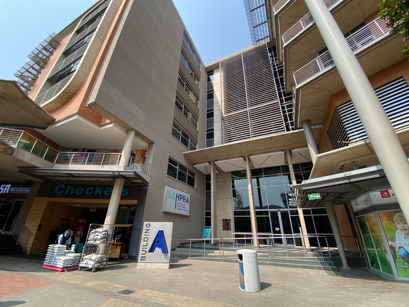 Modern commercial office building entrance featuring a multi-story glass facade, concrete structural elements, and a paved plaza area.
