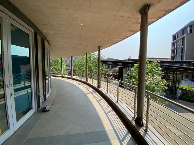 A spacious, curved covered balcony featuring tiled flooring, stainless steel balustrades, and steel support columns, offering an elevated view of the surrounding commercial area.