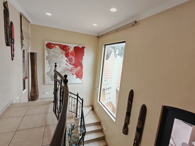 A bright, tiled hallway featuring a wrought iron staircase balustrade, recessed ceiling lighting, and a large window providing natural light.