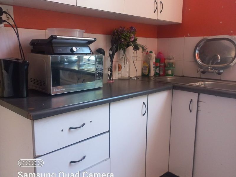A compact kitchen featuring white cabinetry, dark laminate countertops, a stainless steel sink, and a tiled backsplash, currently showing signs of age and wear.
