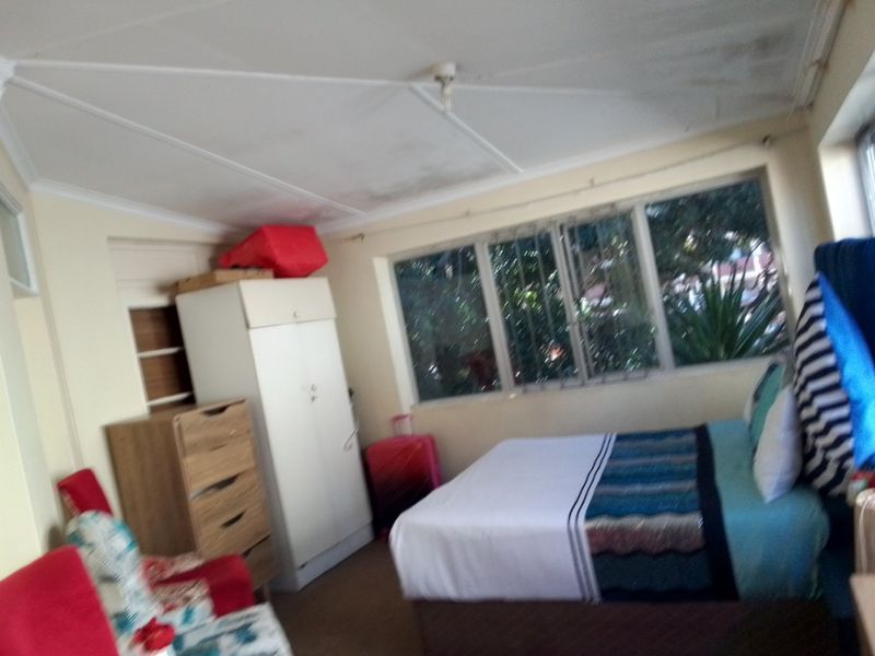 A bedroom featuring a built-in cupboard and large windows with security bars, showing signs of ceiling water damage and wear.