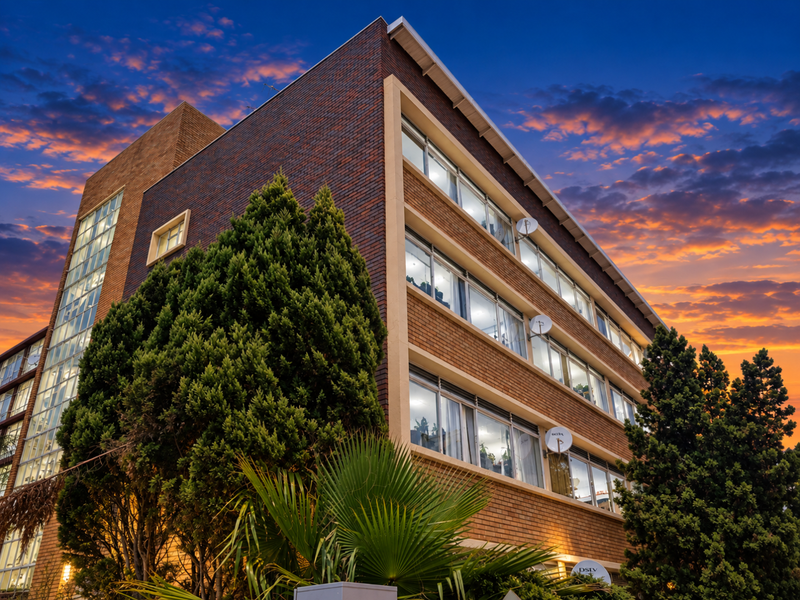 A multi-story commercial building featuring a brick facade, extensive window glazing, and mature landscaping, captured during a vibrant sunset.