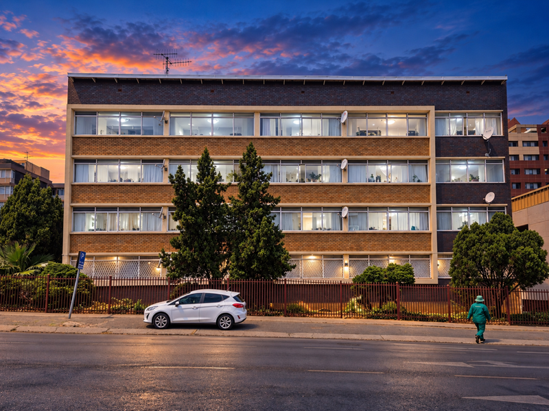 A multi-story commercial or residential building featuring a brick facade, large windows, and secure perimeter fencing, captured during sunset.