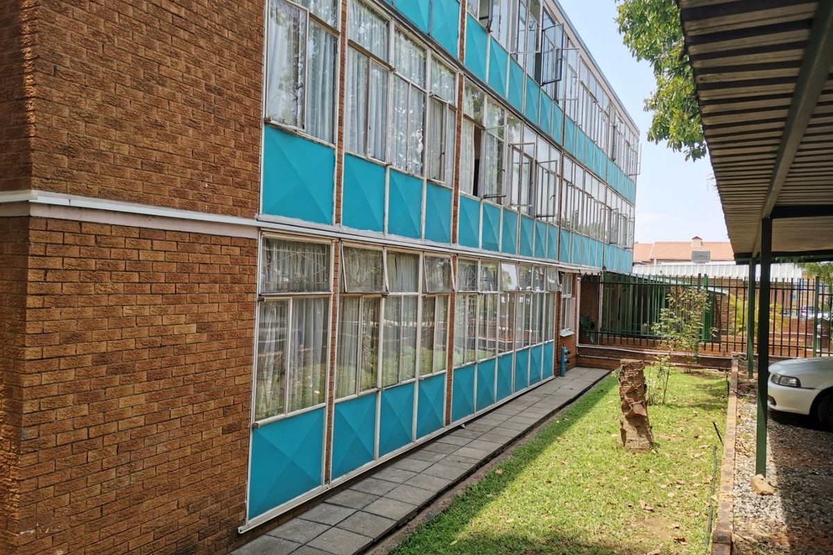 Exterior view of a multi-story commercial building featuring brickwork, blue paneling, and extensive metal-framed windows overlooking a paved walkway and garden area.