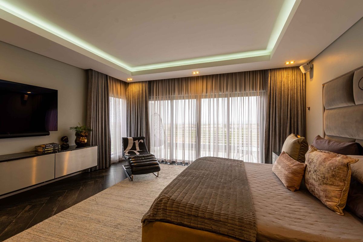 A modern, well-appointed bedroom featuring recessed cove lighting, herringbone timber flooring, and large sliding glass doors leading to an outdoor area.