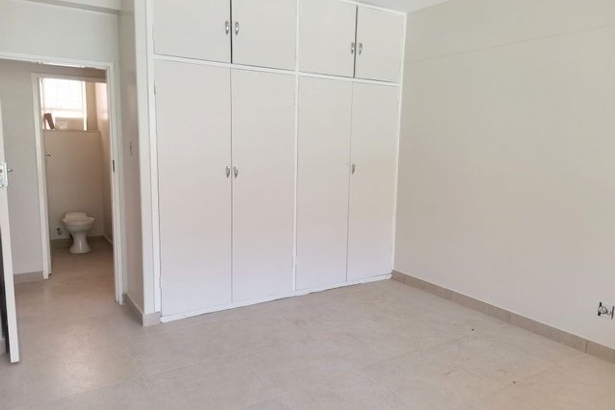 An empty bedroom featuring neutral tiled flooring and extensive built-in cupboard storage, with direct access to an adjoining en-suite bathroom.