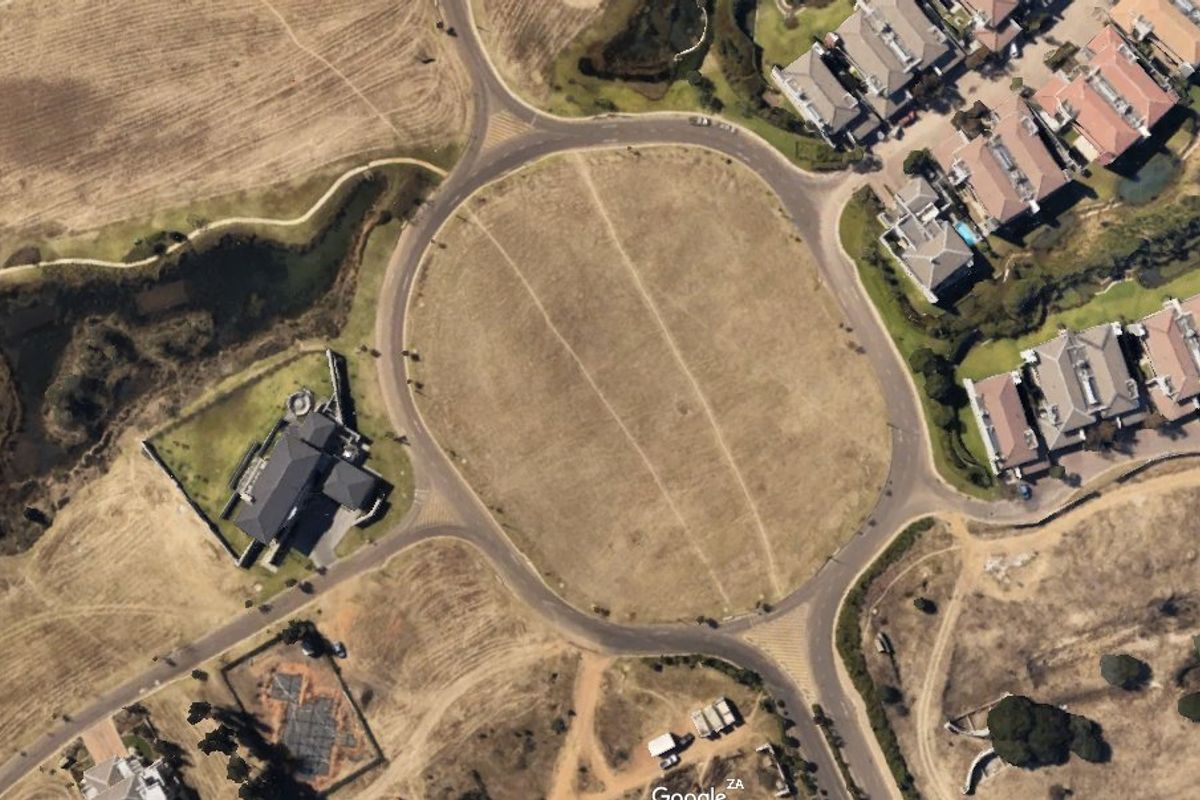 An aerial view of a vacant residential plot situated within a developed estate, surrounded by paved roads and existing residential properties.