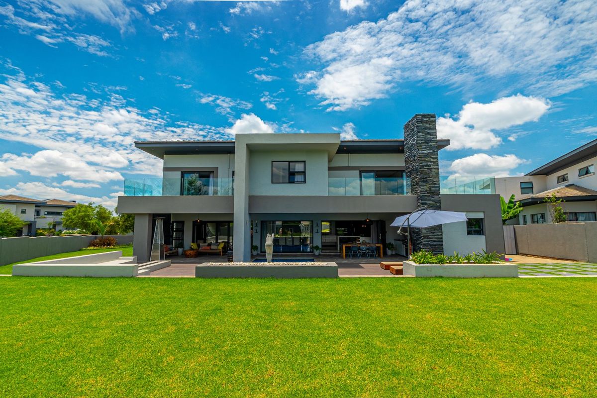 Modern double-story residence featuring a spacious rear patio, stone-clad chimney, glass balustrades, and a well-maintained lawn.